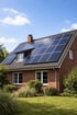 Ziegelrotes Haus mit Solarmodulen auf dem Dach, umgeben von grünen Sträuchern und Rasen unter blauem Himmel.