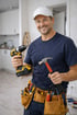 Lächelnder Handwerker mit weißer Kappe, Bohrmaschine, Hammer und Werkzeuggürtel in einer Küche.