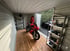Rotes Motorrad in einem offenen Schuppen mit Holzfußboden und Regalen.