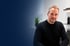Ein lächelnder Mann mit kurzem Haar und Bart im schwarzen Pullover vor einem blau-weiß verlaufenden Hintergrund im Büro.