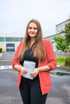 Junge Frau mit langen braunen Haaren, rotem Blazer und schwarzem Oberteil hält eine gerollte Skizze.
