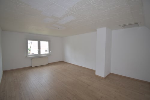 Leerer Raum mit Holzboden, weißen Wänden und einem Fenster mit Heizkörper darunter.