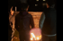Drei Personen stehen um ein brennendes Lagerfeuer im Dunkeln, Holzstapel im Hintergrund.