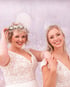 Zwei lächelnde Frauen in weißen Spitzenbrautkleidern vor einem glitzernden, hellvioletten Hintergrund.