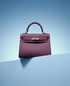 Burgunderrote Handtasche mit Griff und silbernem Verschluss, auf einer blauen Oberfläche vor blauem Hintergrund.