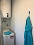 Weißer Warmwasserboiler über einer Waschmaschine mit blauem Handtuch, ein weiteres blaues Handtuch hängt an der Wand.