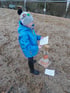 Kind in blauer Jacke und Mario-Mütze hält Papier an einen Pylonen auf einem Reitplatz im Freien.