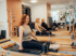 Drei Frauen trainieren in einem hellen Studio auf Pilates-Reformer-Geräten in einer Reihe sitzend.