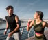 Smiling man and woman jogging outdoors along a river on a sunny day.