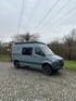Seitenansicht eines Mercedes Sprinter Reisewagens mit Hochdach und Markise – kompakter Ausbau auf Pflasterfläche vor bewaldetem Hintergrund