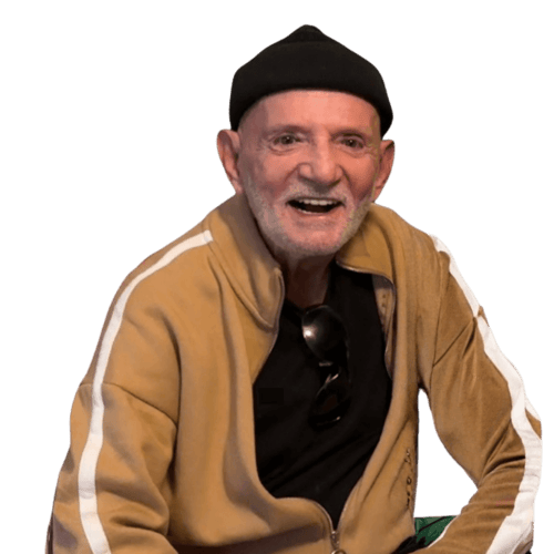 Older man with a beard, black beanie and beige tracksuit, laughing at the camera.