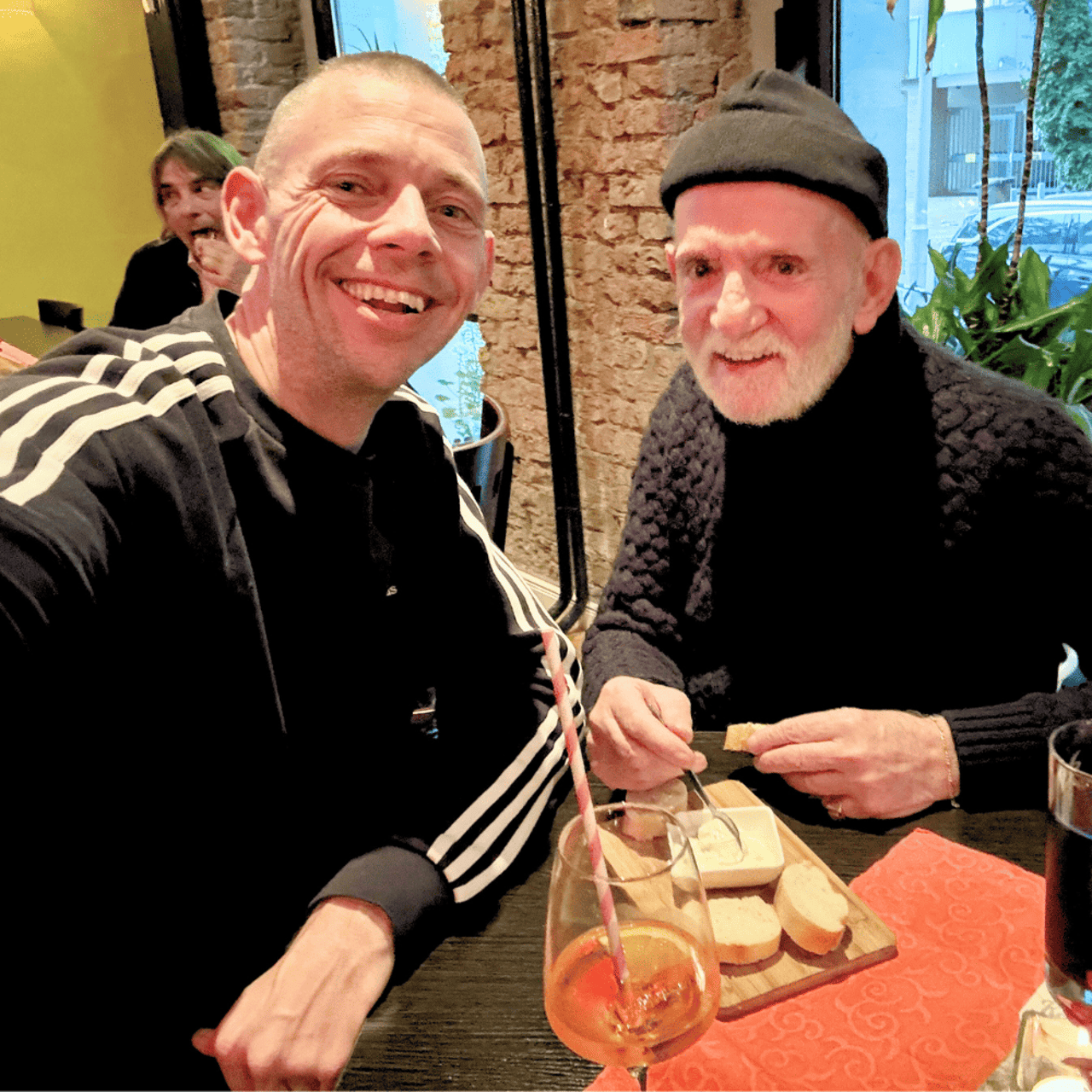 Two men smile at the camera, one with a cap and beard, the other in a black Adidas sweater.