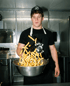 Ein Koch wirft goldgelbe Pommes frites in einer Metallschüssel hoch, während er eine Schürze von Paul's Pommes trägt.