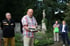 Ein Mann in kariertem Hemd liest aus einem Buch vor einer Gruppe von vier Personen auf einem Friedhof.
