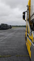 Gelber Lastwagen und eine Reihe militärgrüner Lastwagen auf einem Flugplatz unter bewölktem Himmel.