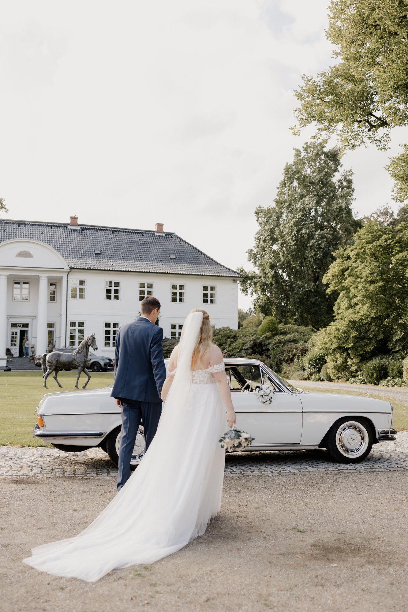Brautpaar geht gemeinsam zum Oldtimer vor dem Gutshaus