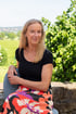 Porträt einer lächelnden Frau mit blonden Haaren im Freien vor einer grünen Hecke und einer weiten Landschaft.