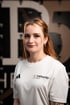 Porträt einer lächelnden Frau mit rötlichem Haar im weißen Sportshirt mit der Aufschrift „THERAPIER this is PHYSIO“ und Pier5.