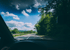 Blick aus dem Autoinnenraum auf eine asphaltierte Landstraße, die an grünen Bäumen unter blauem Wolkenhimmel vorbeiführt.