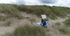 Eine Frau in blauem Pullover sitzt auf einer Decke in den Dünen und malt auf einer Leinwand.