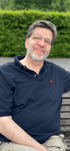 Ein lächelnder Mann mit Bart und Brille in einem marineblauen Poloshirt sitzt vor einer grünen Hecke.