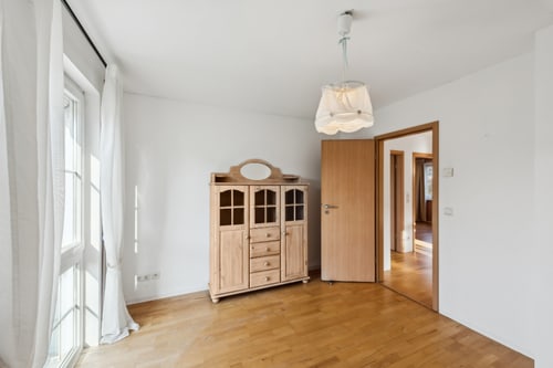 Leeres Zimmer mit Holzboden, weiß gestrichenen Wänden, Fenster, Vorhängen, Holzschrank und offener Tür.