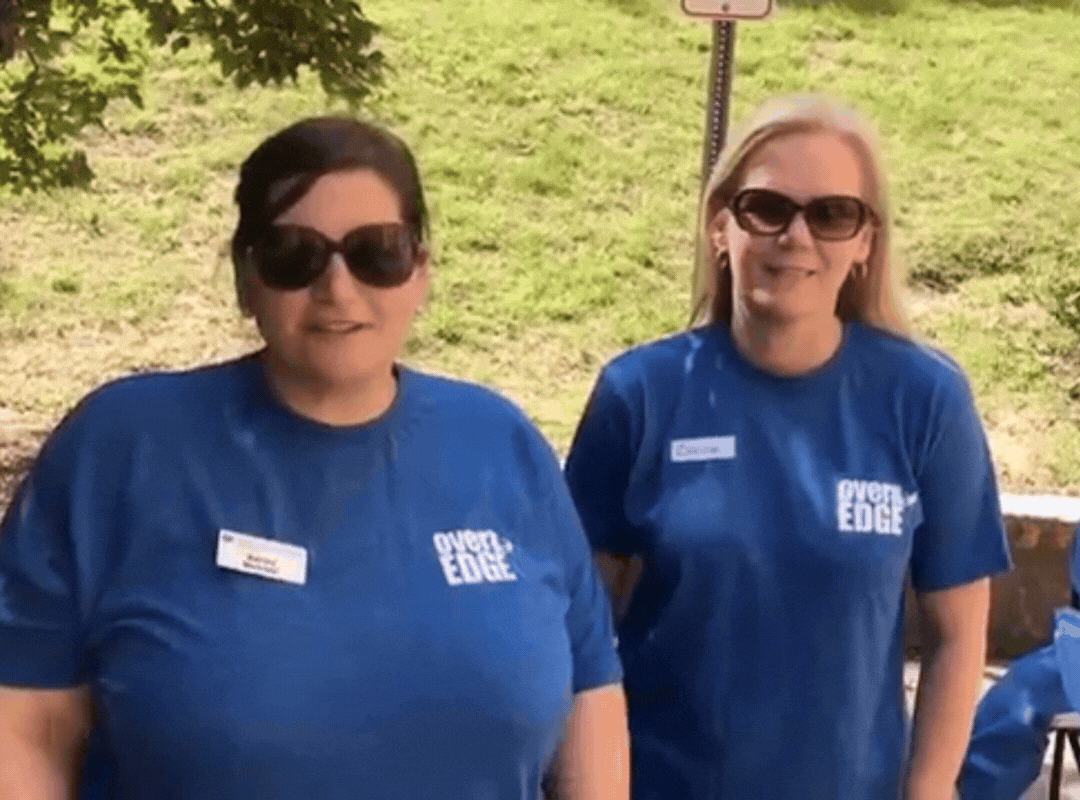 Two women in sunglasses and blue "Over the Edge" event T-shirts with name tags, standing outdoors on a sunny day