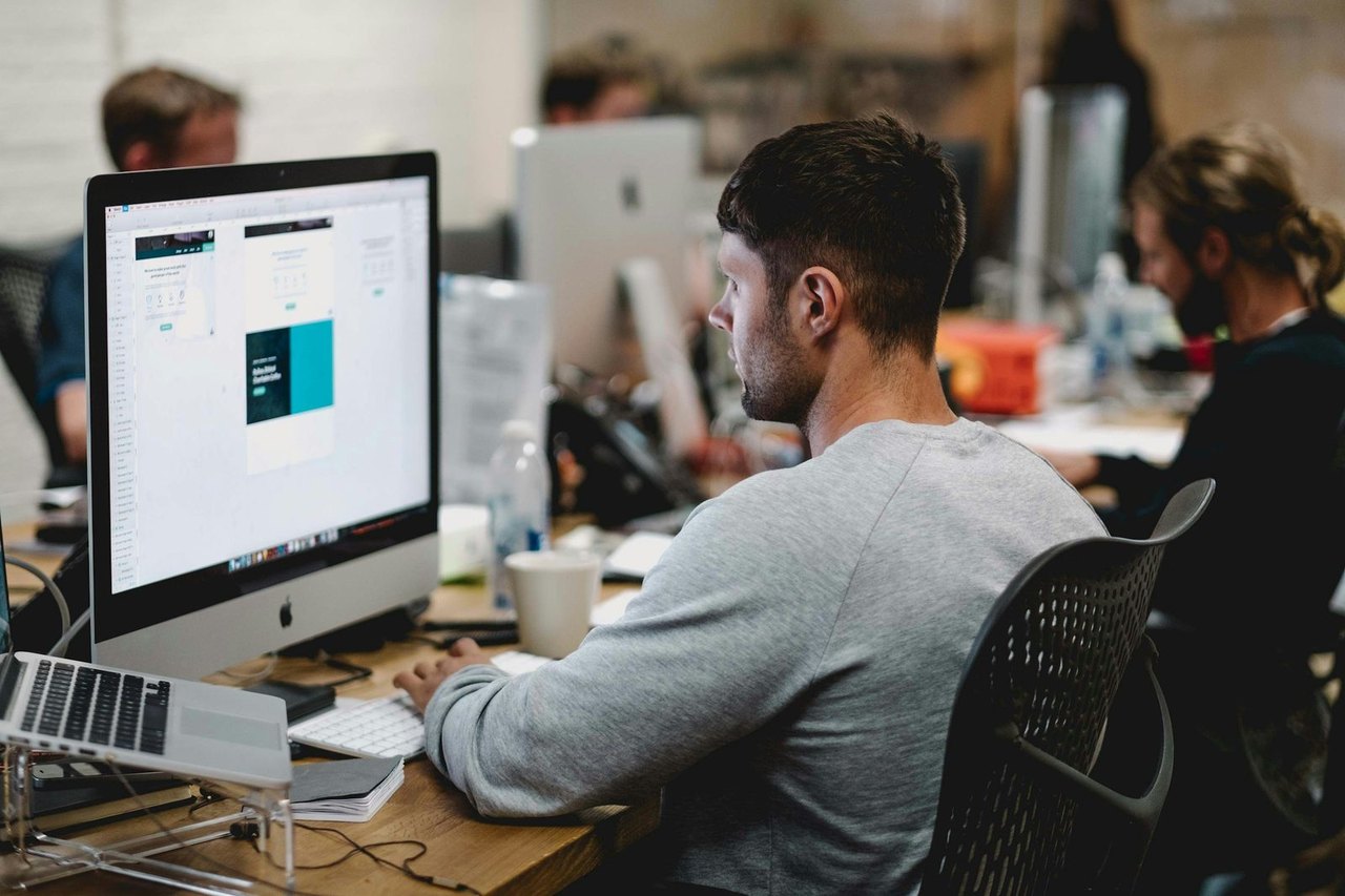 Gestalter arbeitet an einem iMac in einem offenen Agenturbüro, daneben Kollegen