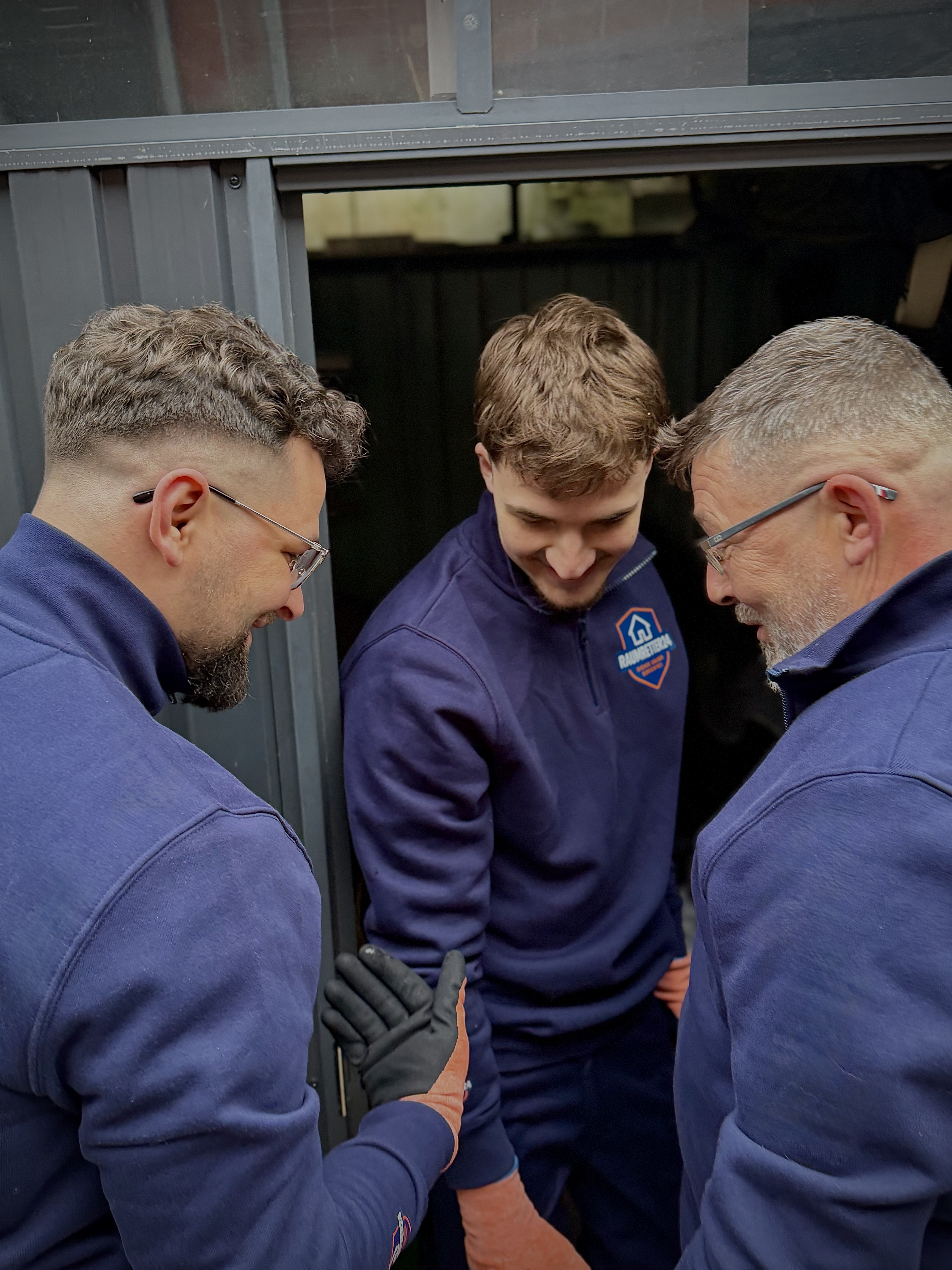 Ein lächelnder Techniker im Blaumann hält ein Klemmbrett und spricht mit einem Kunden auf einer Couch.