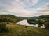 A wide river flows through a valley of lush green hills and forests under a cloudy sky at dusk.