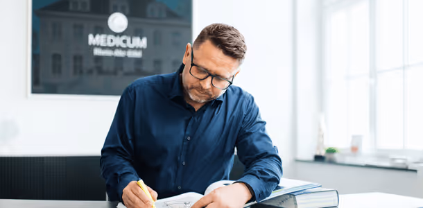 Mann mit Brille in dunkelblauem Hemd schreibt mit gelbem Stift in ein Notizbuch.