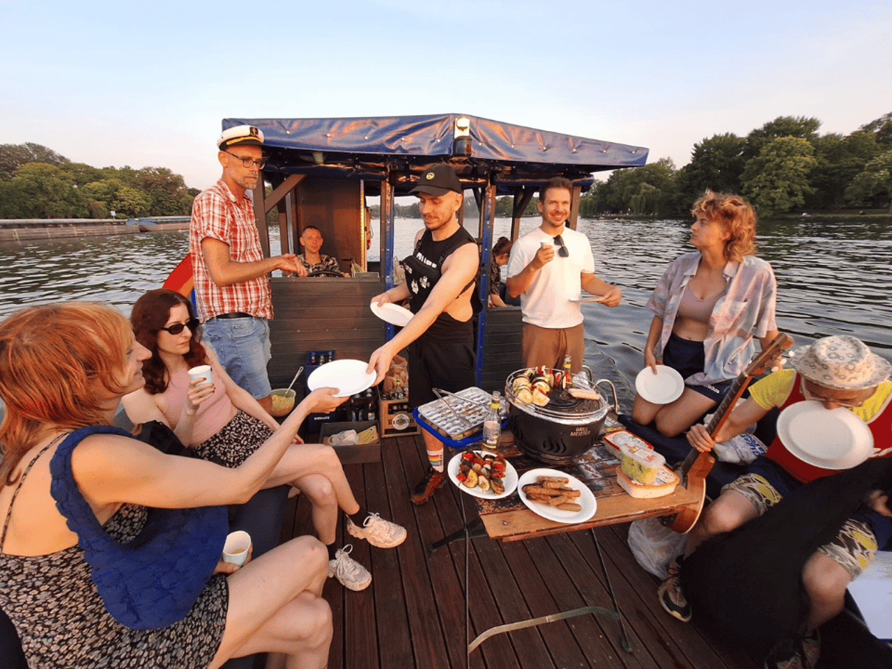 Gruppe von acht Personen grillt und feiert auf einem Pontonboot auf einem See bei Sonnenuntergang.