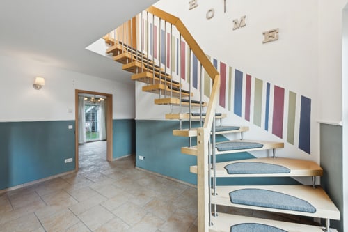 Holztreppe mit Metallgeländer, blauen Teppichstufen und bunten Streifen an der Wand.