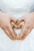 Hände formen ein Herz über einem weißen Hochzeitskleid mit Spitze.