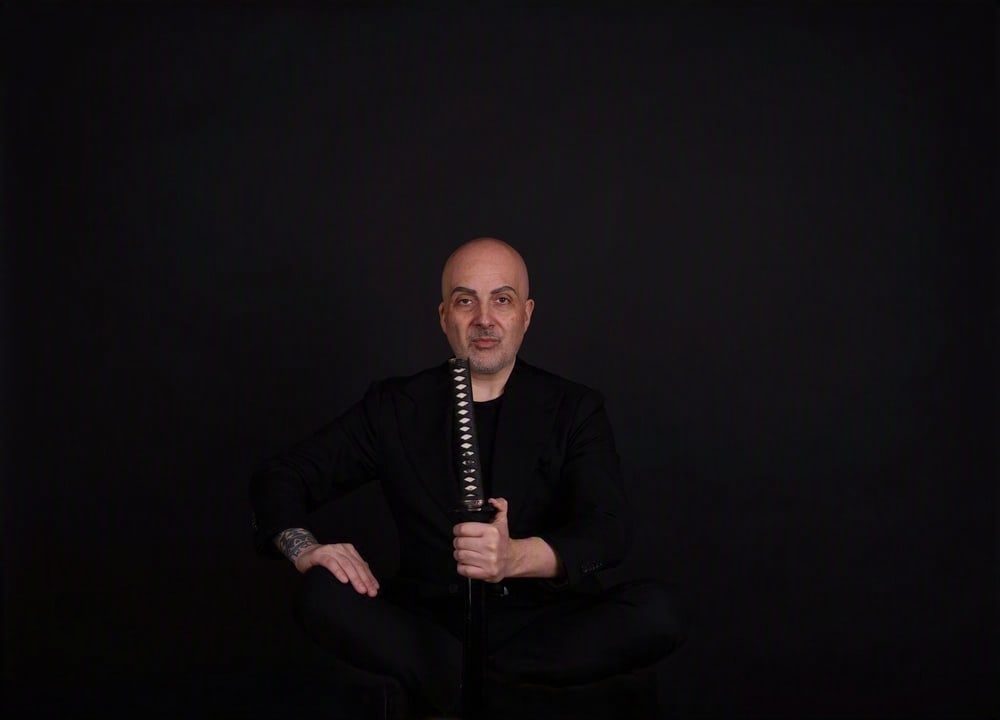 Antonio Bene in Schwarz mit Katana vor dunklem Hintergrund