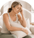 A mature woman sits on a sofa with her eyes closed and hand on her forehead, appearing to have a headache or migraine.