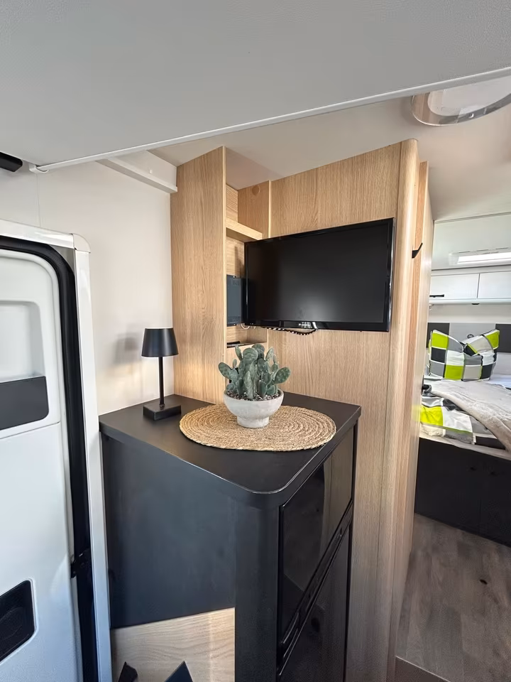 Innenansicht eines Wohnmobils mit schwarzem Kühlschrank, Fernseher, Kaktus und Bett im Hintergrund.