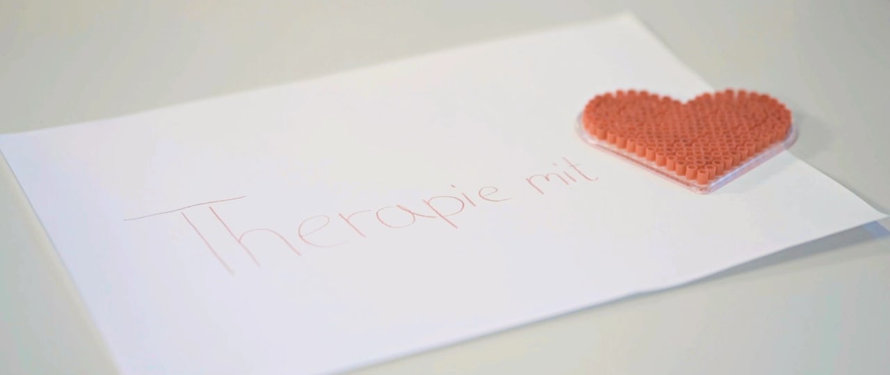 Schriftzug „Therapie mit“ neben einem roten Herz aus Bügelperlen