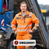 Ein Mann in oranger Arbeitskleidung und Schutzweste steht vor einem blauen Lastwagen.