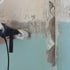 Hand mit Schutzhandschuh bedient einen Bohrhammer an einer blau-weißen Wand.