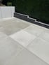 Moderne Terrasse mit großformatigen, hellgrauen Betonplatten vor einer dunklen Steinmauer und einer grünen Hecke.