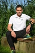 Ein Mann im weißen Poloshirt mit „JENNY PERSONAL TRAINER“ sitzt auf einer Steinmauer vor grünem Schilf.