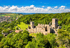 Mittelalterliche Burgruine auf einem bewaldeten Hügel mit einer Stadt im Tal und einem See am Horizont unter blauem Himmel.