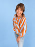 Lächelnde Frau mit braunen Haaren in einer orange-weiß gestreiften Bluse und Jeans vor blauem Hintergrund.