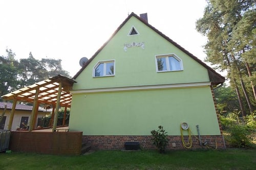 Grünes Haus mit Giebel, zwei Fenstern und gelbem Gartenschlauch; links ein Holzpavillon.