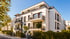 Modernes weißes Mehrfamilienhaus mit Balkonen, Dachterrasse und Hecken unter blauem Himmel.