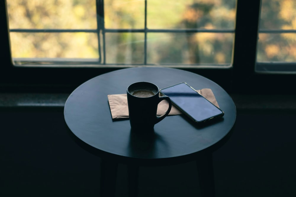 Eine Tasse und ein Smartphone auf einem runden Tisch vor einem Fenster im Halbdunkel