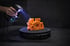 Eine Hand hält einen 3D-Scanner mit blauem Licht, das ein orangefarbenes Bauteil auf einem Drehteller abtastet.