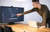 Mann zeigt auf Code auf einem Computermonitor mit einem Laptop auf einem Holztisch.