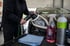 Mann poliert einen Autoscheinwerfer mit einer Poliermaschine; Reinigungsmittel und Tuch liegen daneben.